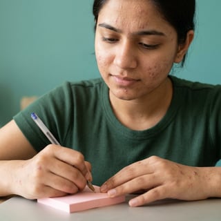 Woman in green shirt writing on pink sticky note at desk against turquoise background